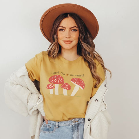 boho girl wearing cute retro mustard colored tee with red and white spotted magic mushrooms with wording above the mushrooms that says "expand your consciousness"